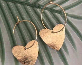 May include: A pair of gold-toned hoop earrings, each featuring a flat, heart-shaped pendant with a brushed metal texture. The circular hoops are thin and metallic, with the heart charms suspended from the bottom of each hoop structure.