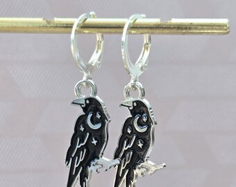 May include: A pair of silver-toned hoop earrings featuring dangling raven charms. Each raven is finished in black enamel with silver-toned celestial details, including a crescent moon and small stars etched into the body of the bird.