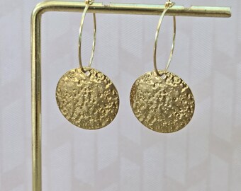 May include: A pair of gold hoop earrings featuring circular, textured pendants. Each earring consists of a thin, polished gold wire hoop that suspends a flat, round disc with a hammered, uneven surface finish that reflects light across its surface.