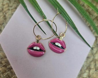 May include: A pair of gold-tone hoop earrings featuring dangling charms shaped like open lips. The lip charms are finished with pink enamel, black detailing, and white accents representing teeth, suspended from thin circular metal hoops.