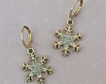 May include: A pair of gold-toned hoop earrings featuring dangling snowflake charms. Each snowflake charm is accented with small, clear, round-cut crystals embedded in the center, creating a sparkling effect on the metallic snowflake design.