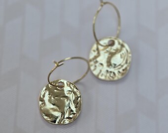 May include: A pair of gold hoop earrings featuring circular, hammered-texture charms. Each thin gold hoop suspends a round, reflective metal disc with an uneven, organic surface finish. The earrings are polished and catch the light, showing a bright, metallic gold tone throughout the entire design.