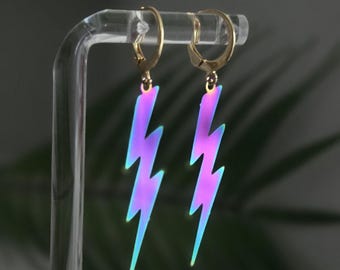 May include: A pair of gold-toned hoop earrings featuring dangling lightning bolt charms. The lightning bolts have an iridescent finish that shifts between vibrant shades of purple, pink, and blue, creating a reflective and colorful appearance.