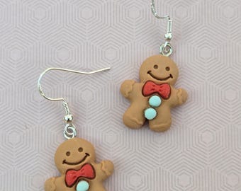 May include: A pair of dangle earrings featuring gingerbread man charms. Each charm is light brown with a smiling face, a red bow tie, and two light blue circular buttons on the torso. The charms hang from silver-toned metal fishhook ear wires.