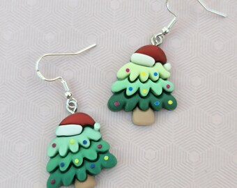 May include: A pair of dangle earrings featuring Christmas tree charms with light and dark green layers, a brown trunk, and a red Santa hat on top. The trees are decorated with small, colorful dots and are attached to silver-toned metal earring hooks.