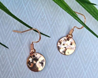May include: A pair of rose gold-toned drop earrings featuring small, circular, hammered metal discs. Each earring is attached to a simple curved ear wire hook, showcasing a reflective, textured surface that catches the light with its uneven finish.
