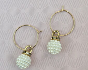 May include: A pair of gold hoop earrings featuring a dangling charm composed of a cluster of small white faux pearls. Each charm is accented with a thin gold band embedded with tiny clear crystals at the top of the pearl cluster.