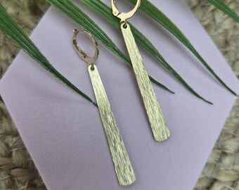 May include: A pair of gold-toned drop earrings featuring a brushed metal finish. Each earring consists of a circular hoop closure attached to a long, slender, rectangular bar pendant with a textured surface that catches the light.