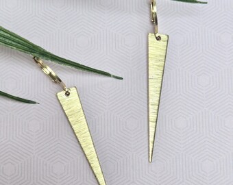 May include: A pair of gold-toned earrings featuring long, slender, triangular pendants with a brushed metal texture. Each pendant is suspended from a small, circular hoop closure, creating a clean, geometric design with a metallic finish.