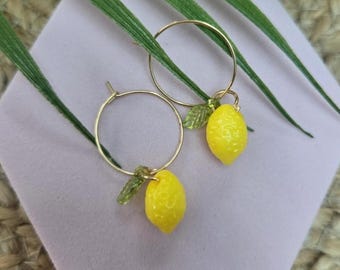 May include: A pair of gold-toned hoop earrings featuring small, bright yellow lemon charms and tiny, translucent green leaves attached to each hoop. The earrings are displayed on a light purple hexagonal base with green foliage visible.