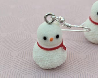 May include: A pair of dangle earrings featuring white snowman charms with textured surfaces, small black eyes, orange carrot-shaped noses, and thin red scarves. The charms are attached to silver-toned metal fishhook ear wires for hanging.