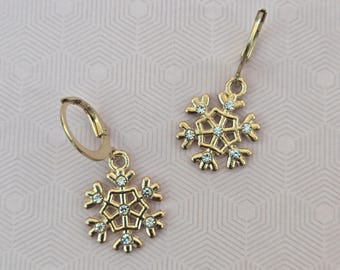 May include: A pair of gold-toned hoop earrings featuring dangling snowflake-shaped charms. Each snowflake charm is crafted with a geometric openwork design and is accented with small, clear, sparkling crystals embedded in the metal frame.