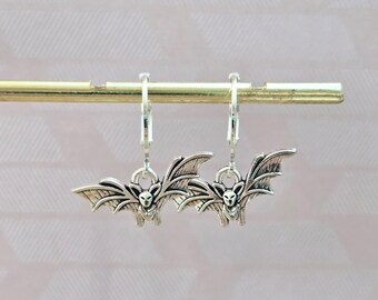 May include: A pair of silver-toned metal hoop earrings featuring bat-shaped charms. Each charm depicts a detailed bat with spread wings and pointed ears, suspended from a small circular hoop mechanism designed for attachment to the earlobe.