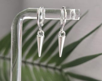 May include: A pair of silver-colored hoop earrings with a conical, pointed charm hanging from each hoop. The earrings are displayed on a clear acrylic stand against a blurred background of green leaves.