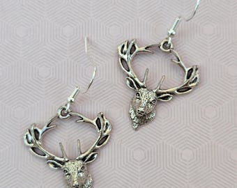 May include: A pair of silver-toned drop earrings featuring a detailed deer head design with prominent antlers forming a circular frame. Each earring includes a thin metal hook for attachment, showcasing a metallic finish with dark recessed details.