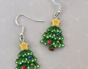May include: A pair of dangle earrings featuring green Christmas tree charms with brown trunks, yellow star toppers, and small colorful ornaments. The trees are decorated with white dots and are attached to silver-toned metal fishhook ear wires.