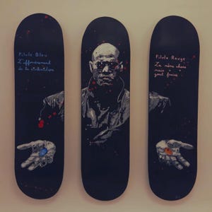 May include: Three black skateboards with artwork. The centre board features a portrait of a man in sunglasses. The other two boards have text and a hand holding a pill. One says "Pilule Bleu" and the other says "Pilule Rouge".