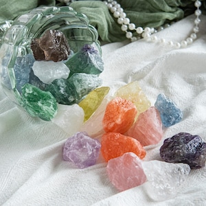 Peut inclure: Un bol en verre transparent renversé, déversant une variété de pierres précieuses brutes colorées. Les pierres sont dans des tons de vert, violet, orange, rose, jaune, bleu et transparent. Le bol est posé sur un tissu blanc texturé, avec un collier de perles et un tissu vert en arrière-plan.