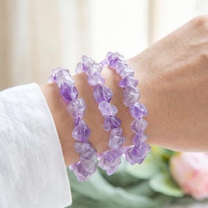 Puede incluir: Se muestran tres pulseras de virutas de amatista en una muñeca. Las pulseras están hechas de piedras irregulares de color púrpura claro ensartadas. La persona lleva una camisa blanca de manga larga. El fondo está borroso, con un toque de flor rosa.