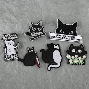 May include: A collection of embroidered cat patches in various designs. One patch features a white cat with the text "I AM SMALL AND SENSITIVE BUT ALSO FIGHT ME." Other patches include a black cat with a knife, and a cat with the text "DON'T GO OUTSIDE, THERE ARE PEOPLE-OUT THERE."