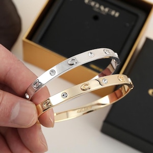 May include: Two hinged bangle bracelets, one in silver tone and one in gold tone, each featuring alternating clear crystal accents and small metal C-shaped logos along the outer surface, presented with a black Coach branded jewellery box.