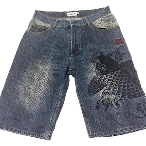May include: Blue denim shorts featuring a dark wash with a subtle snakeskin pattern throughout. The front includes a large black embroidered koi fish design on the right leg, a small red square patch, and light grey snakeskin-textured pockets.