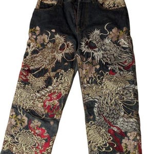 May include: Dark wash denim jeans featuring detailed embroidery of dragons and floral motifs in gold, red, and cream. The embroidery adorns the front of the legs. The jeans have a classic five-pocket design. The measurements are in centimeters.