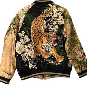以下が含まれることがあります： 黒と金のサテン製ボンバージャケット。背中には、白い桜の花と緑の竹に囲まれた虎の精巧な刺繍が施されています。このジャケットは、長い金色の袖、リブ編みの襟、そして黒いストライプが入ったリブ編みの袖口が特徴です。