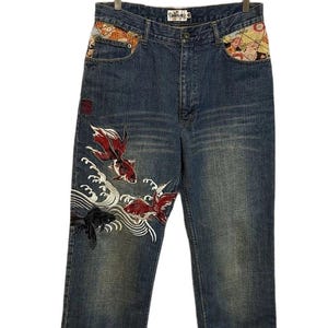 Peut inclure: Jean en denim bleu à taille haute avec des empiècements en tissu à motifs colorés au niveau des poches latérales. La jambe droite présente une broderie détaillée de deux poissons rouges et d'un poisson noir nageant parmi des vagues blanches.