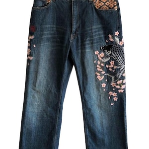 May include: Dark blue denim jeans featuring an embroidered black and white koi fish surrounded by pink cherry blossoms on the left leg. The waistband interior and front pocket linings are accented with a tan and brown geometric patterned fabric.