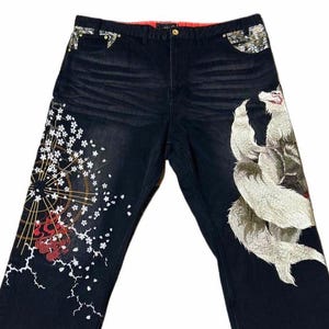 Può includere: Jeans in denim blu scuro con ricami elaborati. I jeans presentano un grande lupo ricamato bianco e grigio su una gamba e un design floreale e a fulmini sull'altra. Le tasche hanno tessuto decorativo.