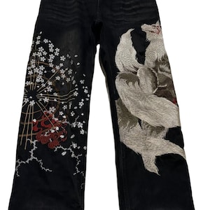 May include: Black denim jeans featuring an embroidered white multi-tailed fox on the right leg and a large circular pattern with white cherry blossoms and red accents on the left leg. The waistband has patterned fabric inserts at the front pockets.