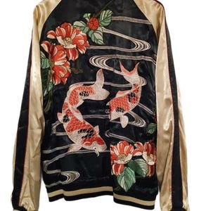 Könnte beinhalten: Eine schwarze Satin-Bomberjacke mit goldenen Ärmeln und gerippten Bündchen. Die Rückseite zeigt eine detaillierte Stickerei von zwei Koi-Fischen, die zwischen stilisierten Wasserströmungen schwimmen, akzentuiert durch große rote und grüne Blüten.