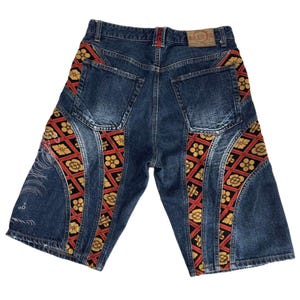 May include: Dark blue denim shorts featuring two back pockets and panels of black fabric with a red grid pattern and gold floral motifs. The shorts include a tan leather brand patch on the waistband and white line art on the left leg panel.