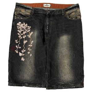 May include: Black denim shorts featuring a faded wash and a textured, crinkled appearance. The front displays an embroidered branch of light pink cherry blossoms on the left side, with dark patterned fabric accents on the pockets and a brown waistband.