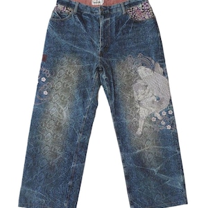 May include: Blue acid-wash denim jeans featuring a large embroidered koi fish and floral patterns on the right leg. The front pockets are lined with a dark fabric featuring a small floral print. The waistband interior is a solid muted red colour.
