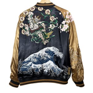 Puede incluir: Una chaqueta bomber de satén negro con mangas doradas que presenta un intrincado bordado en la espalda. El diseño muestra a un dios del trueno rodeado de tambores, una gran ola azul y flores de cerezo rosas. El cuello, puños y bajo son acanalados.