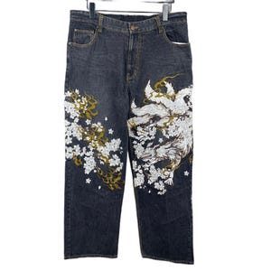 May include: Dark grey denim jeans featuring intricate white and gold embroidery. The design includes a large multi-tailed fox figure on the right leg and a dense pattern of cherry blossom flowers and swirling gold accents across both legs.