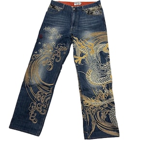 Könnte beinhalten: Blaue Jeans aus Denim mit aufwendigen gold- und weißfarbenen Stickereien eines großen Drachen und wirbelnden Wellenmustern auf den Beinen. Der Bund ist mit dekorativen schwarzen und silbernen Blumenstickereien an den Vordertaschen und einem orangefarbenen Innenfutter versehen.