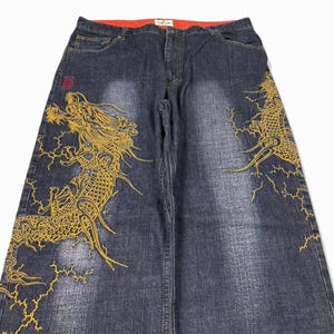 Op de afbeelding: Donkerblauwe denim jeans met een rode binnenband. De jeans hebben een grote gouden drakenborduurontwerp op de voor- en zijkanten. Het drakenontwerp is gedetailleerd met bliksemaccenten.