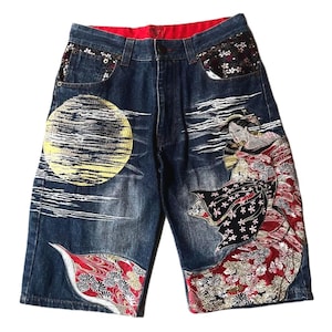 May include: Dark blue denim shorts featuring an embroidered yellow full moon on the left leg and a geisha figure in a red and black floral kimono on the right leg. The pockets have black floral fabric accents and the interior waistband is red.