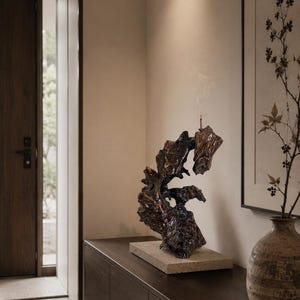 May include: A dark brown, organic-shaped wooden sculpture with a glossy finish, mounted on a rectangular stone base. A thin incense stick is inserted into the top of the wood, emitting a faint trail of smoke into the air above the sculpture.