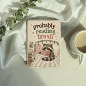 May include: A rectangular wooden sign featuring a raccoon wearing glasses and reading a book in bed. The text reads, "probably reading trash." Beside the raccoon is a stack of books and a potted plant. The sign has a light cream background.