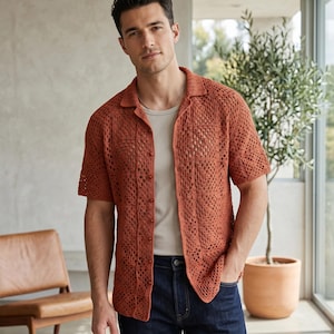 May include: A short-sleeved, open-weave crochet shirt in a warm terracotta color. The shirt has a collared neckline and button-down front, paired with a light-colored undershirt and dark blue jeans. The shirt's open weave design is visible.