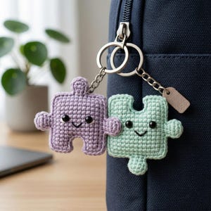 May include: A pair of crocheted puzzle piece keyrings in lavender and mint green, each with a smiling face. They are attached to a silver keyring and a navy blue bag.