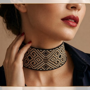 Puede incluir: Un collar gargantilla de cuentas negras y doradas con un patrón geométrico. La gargantilla ancha se coloca alrededor del cuello, mostrando un diseño detallado. La modelo lleva una blusa azul oscuro.