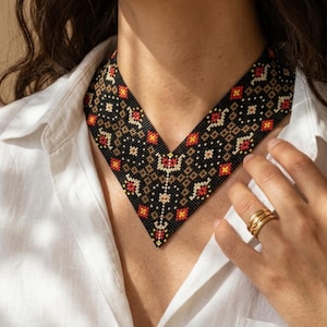 May include: A black beaded necklace with a geometric pattern in red, yellow, and tan. The necklace is worn over a white collared shirt. The design is a V-shape, and the beads are small and tightly woven.