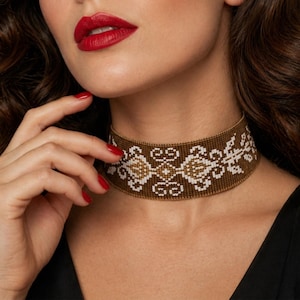 May include: A close-up of a brown beaded choker necklace with white and gold geometric patterns. The necklace is worn around the neck. The person in the image has red lipstick and red nail polish.
