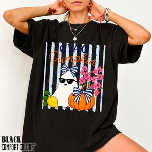 May include: A black short-sleeve t-shirt featuring a graphic print of a white ghost wearing black sunglasses and a blue and white striped bow. Beside the ghost is an orange pumpkin with a matching bow, a yellow lemon, and pink flowers.