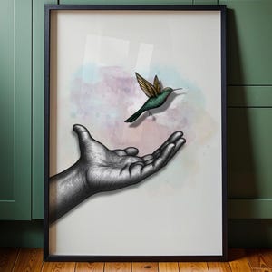 Puede incluir: Un cuadro enmarcado con una mano extendida hacia un colibrí. El colibrí tiene alas verdes y marrones, sobre un fondo de acuarela suave en tonos rosas y azules. La impresión está en un marco negro.